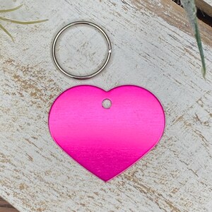 May include: A pink heart-shaped metal tag with a silver ring attached. The tag has a hole in the top for attaching to a key chain or other item.
