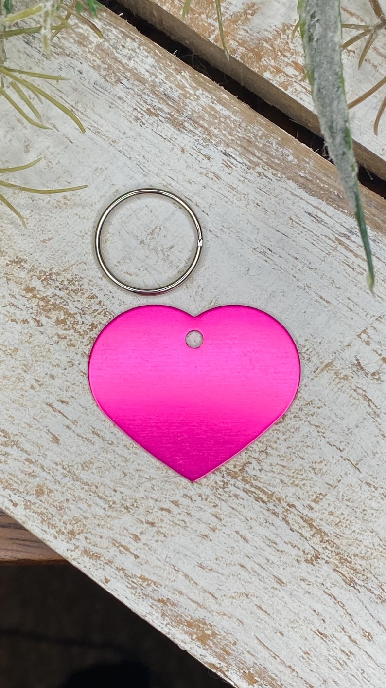 May include: A pink heart-shaped metal tag with a silver ring attached. The tag has a hole in the top for attaching to a key chain or other item.