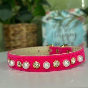 Könnte beinhalten: Ein leuchtend pinkfarbenes Hundehalsband mit weicher, samtiger Textur. Das Halsband ist mit abwechselnden Kunstperlen und klaren, goldfarbenen Strasssteinen verziert. Es hat eine goldfarbene Schnalle.