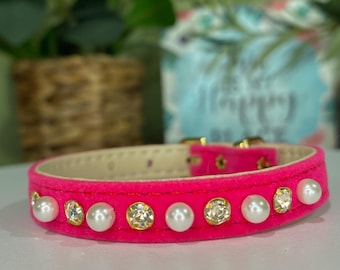 Könnte beinhalten: Ein leuchtend pinkfarbenes Hundehalsband mit weicher, samtiger Textur. Das Halsband ist mit abwechselnden Kunstperlen und klaren, goldfarbenen Strasssteinen verziert. Es hat eine goldfarbene Schnalle.