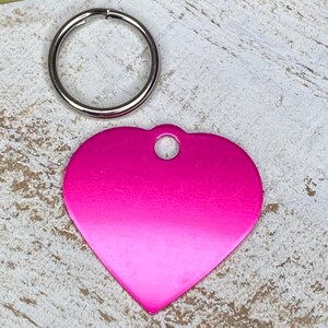 May include: A pink heart-shaped metal tag with a silver ring attached. The tag has a smooth, shiny finish and a hole at the top for attaching to a key chain or collar.