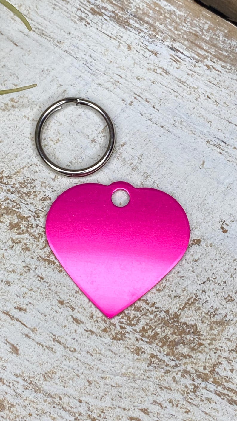 May include: A pink heart-shaped metal tag with a silver ring attached. The tag has a smooth, shiny finish and a hole at the top for attaching to a key chain or collar.