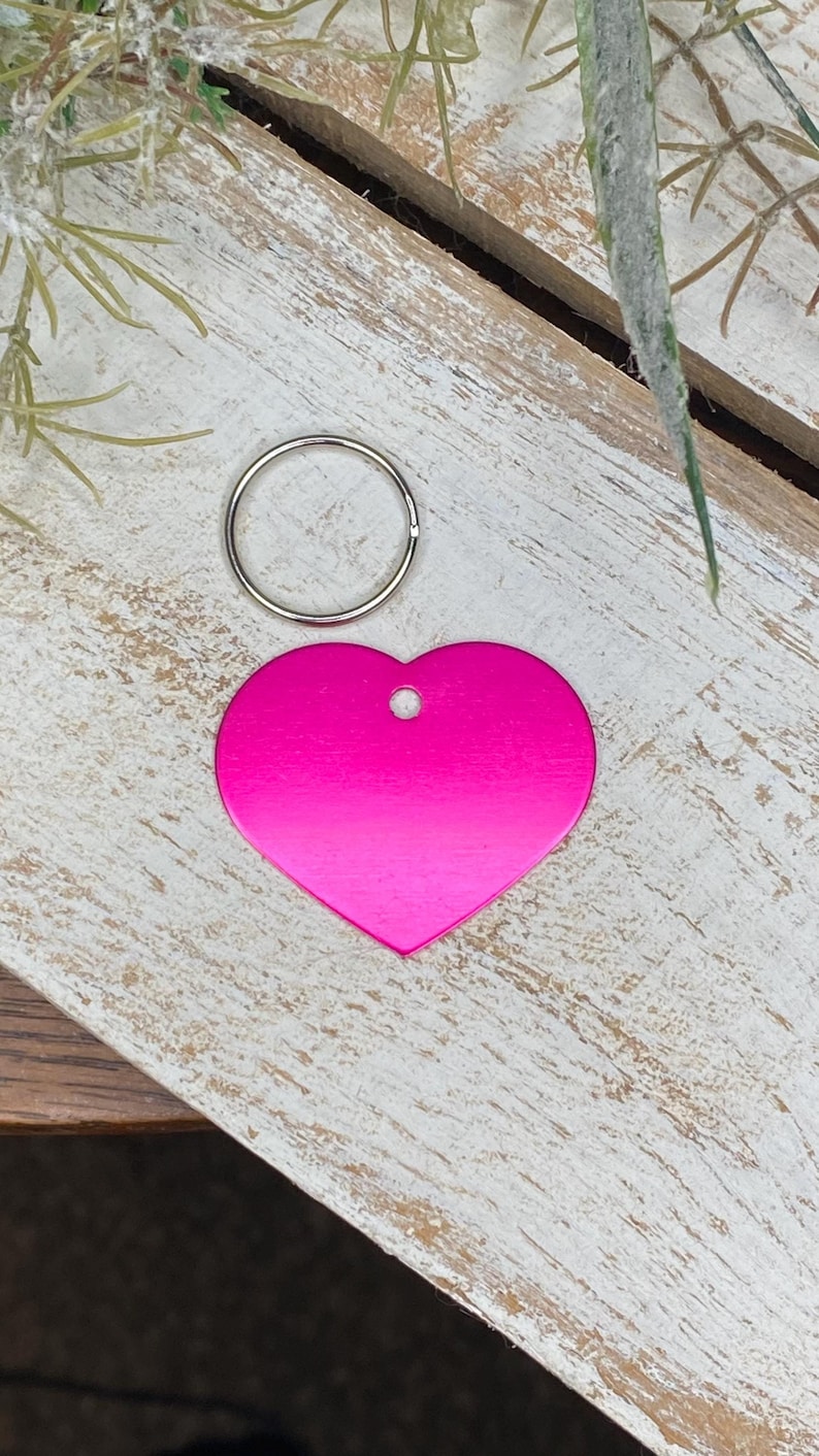 May include: A bright pink heart-shaped metal tag with a silver key ring. The tag has a hole at the top for attaching to a collar or leash.