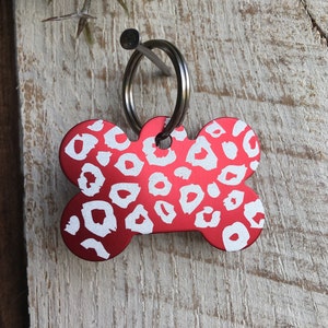 May include: Red bone-shaped dog tag with a white leopard print design. The tag has a silver ring attached.