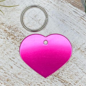 May include: A pink heart-shaped metal tag with a hole in the top for a key ring. The tag is on a white wooden surface.