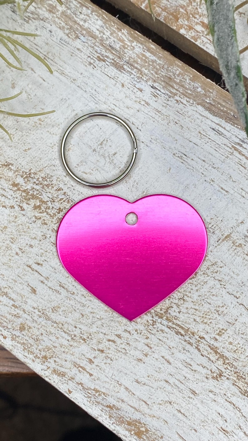 May include: A pink heart-shaped metal tag with a hole in the top for a key ring. The tag is on a white wooden surface.