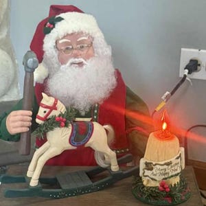 May include: A Christmas-themed Santa Claus figurine with a workshop scene. Santa holds a hammer and stands near a rocking horse. The workshop includes a candle with the text "Merry Christmas and Happy New Year" and wrapped gifts.