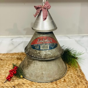 May include: A rustic Christmas tree made from galvanized metal funnels. The tree is topped with a red and white checkered bow. The base is made of two large funnels, with a smaller one on top. The tree is decorated with a sprig of greenery and red berries.