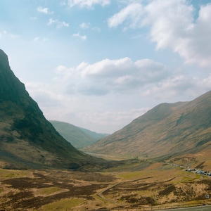 May include: A view of a valley in the Scottish Highlands with two large mountains on either side. The valley is covered in green grass and there are a few cars parked on the road.