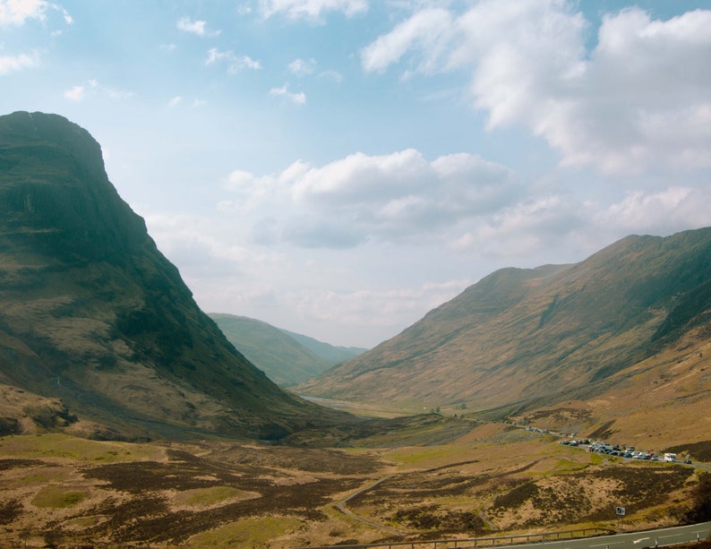 May include: A view of a valley in the Scottish Highlands with two large mountains on either side. The valley is covered in green grass and there are a few cars parked on the road.
