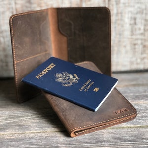 May include: Two brown leather passport holders. The top holder is open and a blue US passport is visible. The bottom holder is closed and has the initials "EAK" embossed on the front.