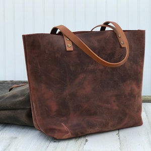 May include: A large, dark brown leather tote bag with a distressed finish and two lighter tan leather shoulder straps attached with metallic rivets. A second, smaller dark grey leather bag is partially visible behind the main tote.