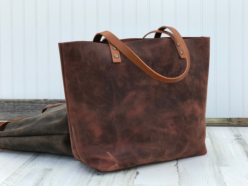 May include: A large, dark brown leather tote bag with a distressed finish and two lighter tan leather shoulder straps attached with metallic rivets. A second, smaller dark grey leather bag is partially visible behind the main tote.