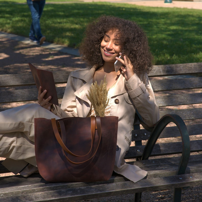 May include: A large, dark brown leather tote bag with two matching shoulder straps. The bag has a smooth, matte finish and a spacious, open-top design. A bundle of dried wheat stalks is placed inside the bag, extending upwards from the opening.