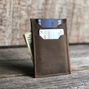 K&ouml;nnte beinhalten: Eine braune Ledergeldb&ouml;rse mit zwei Kartenf&auml;chern und einem Fach f&uuml;r Bargeld. Die Geldb&ouml;rse ist geschlossen und hat einen Used-Look.