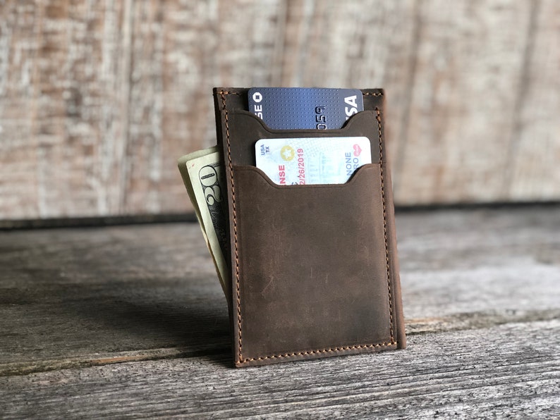 K&ouml;nnte beinhalten: Eine braune Ledergeldb&ouml;rse mit zwei Kartenf&auml;chern und einem Fach f&uuml;r Bargeld. Die Geldb&ouml;rse ist geschlossen und hat einen Used-Look.