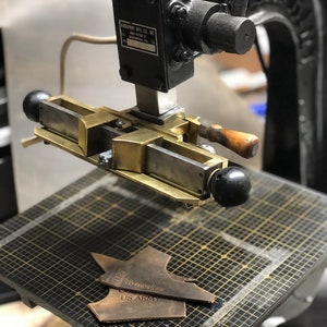 May include: A close-up of a vintage leather stamping machine with a metal base and a lever. The machine is being used to stamp a piece of leather with the text "To my dear US Army".