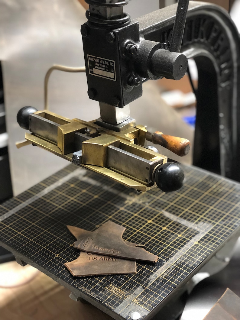 May include: A close-up of a vintage leather stamping machine with a metal base and a lever. The machine is being used to stamp a piece of leather with the text "To my dear US Army".