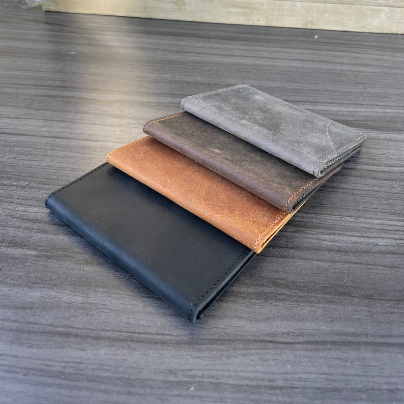 May include: Four leather wallets stacked on top of each other. The top wallet is gray, the second is brown, the third is brown with a lighter shade, and the bottom wallet is black.