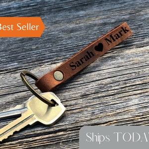 May include: Personalized leather keychain with the names "Sarah" and "Mark" separated by a heart symbol. The keychain is brown and has a metal ring for attaching keys. The text "Color Shown: CINNAMON" is visible in the image.