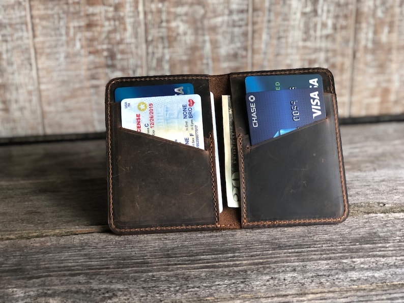May include: A brown leather wallet with a flap closure. The wallet has two card slots and a billfold. The wallet is open and shows a driver's license, a credit card, and cash.