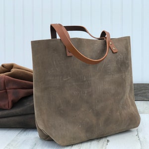May include: A large, taupe-colored leather tote bag with a matte, distressed finish and two tan leather shoulder straps attached with metal rivets. Behind the main bag, two additional folded leather bags in dark brown and tan are visible.