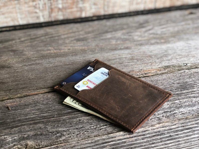K&ouml;nnte beinhalten: Eine braune Ledergeldb&ouml;rse mit abgenutztem Finish. Die Geldb&ouml;rse hat ein Kartenfach und ein Fach f&uuml;r Bargeld. Eine Kreditkarte und Bargeld sind in der Geldb&ouml;rse sichtbar.