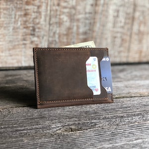 K&ouml;nnte beinhalten: Eine braune Ledergeldb&ouml;rse mit einem Kartenfach und einem Fach f&uuml;r Bargeld. Die Geldb&ouml;rse ist geschlossen und enth&auml;lt eine Kreditkarte und Bargeld.