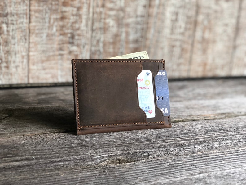 K&ouml;nnte beinhalten: Eine braune Ledergeldb&ouml;rse mit einem Kartenfach und einem Fach f&uuml;r Bargeld. Die Geldb&ouml;rse ist geschlossen und enth&auml;lt eine Kreditkarte und Bargeld.