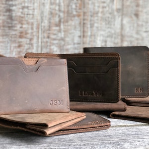 May include: Three brown leather wallets with different personalized engravings. The wallets have multiple card slots and a billfold. The wallets are arranged on a wooden surface.