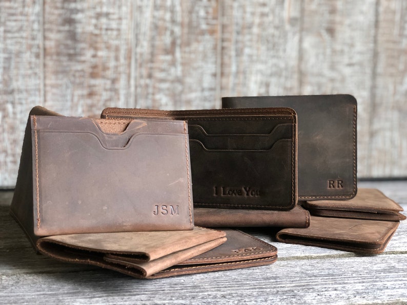 May include: Three brown leather wallets with different personalized engravings. The wallets have multiple card slots and a billfold. The wallets are arranged on a wooden surface.
