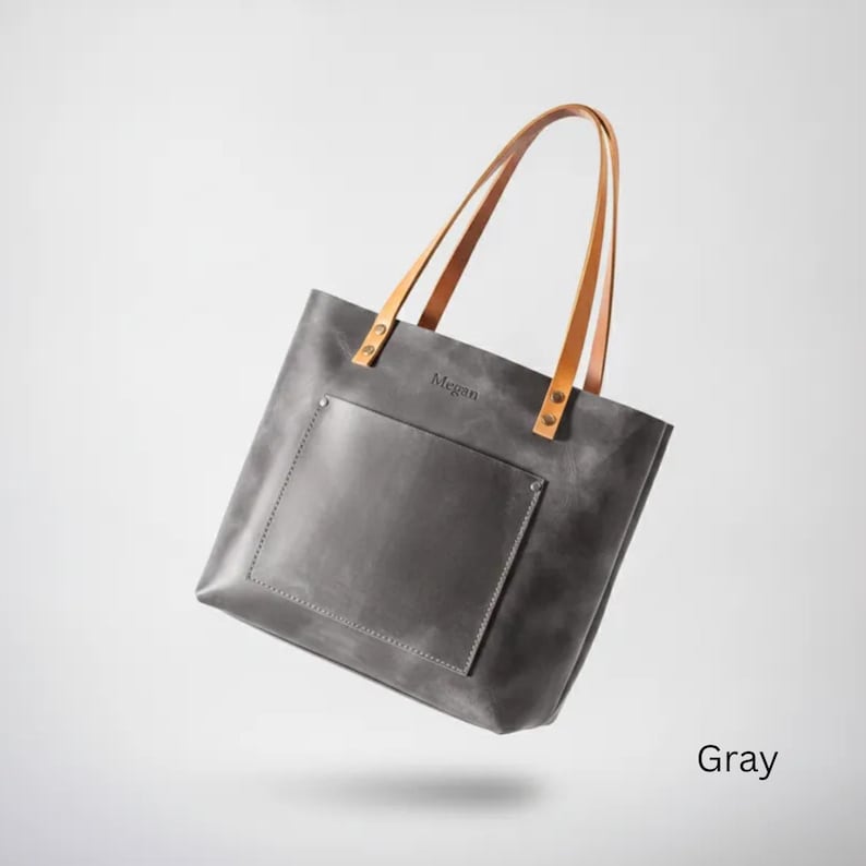 May include: A gray leather tote bag featuring two tan leather shoulder straps attached with metal rivets. The bag has a large front pocket and the name Megan embossed in the center. The design includes visible stitching along the pocket edges.