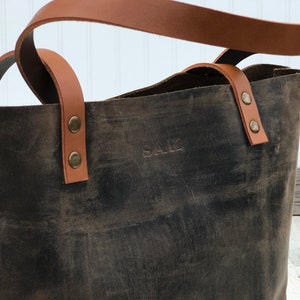May include: A dark brown leather tote bag featuring tan leather handles attached with brass-colored metal rivets. The front of the bag is embossed with the letters SAK in a simple, capitalized font, showcasing a distressed texture throughout.