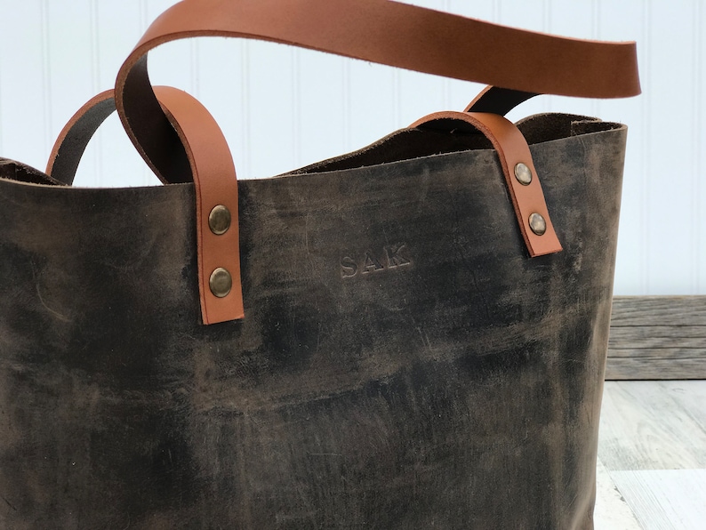 May include: A dark brown leather tote bag featuring tan leather handles attached with brass-colored metal rivets. The front of the bag is embossed with the letters SAK in a simple, capitalized font, showcasing a distressed texture throughout.