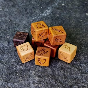 Puede incluir: Un conjunto de dados de madera con símbolos grabados, incluyendo espadas, escudos y rayos. Los dados son de varios tonos de marrón y beige, con un dado marrón oscuro. Están colocados sobre una superficie de piedra gris. El texto "Wood Tactics LLC ©" es visible.