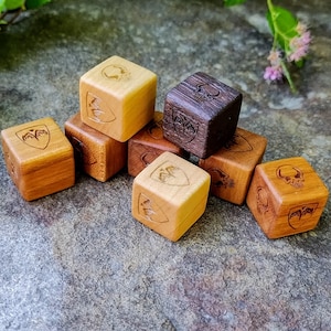 May include: A selection of wooden dice, each featuring engraved designs such as skulls and crests. The dice are in shades of brown and tan. The text "Wood Tactics LLC ©" is visible.