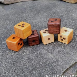 May include: A collection of wooden dice in various shades of brown and tan, each with three inlaid dark squares. The dice are arranged on a gray stone surface, with natural stone elements in the background. The text "Wood Tactics LLC ©" is visible.