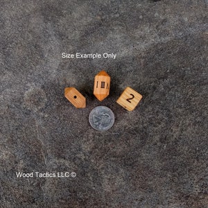 Puede incluir: Cuatro dados de madera con diferentes n&uacute;meros y s&iacute;mbolos en cada cara. Los dados est&aacute;n dispuestos en c&iacute;rculo sobre una superficie gris texturizada. El texto "Size Example Only" es visible sobre los dados. El texto "Wood Tactics LLC &copy;" es visible debajo de los dados.