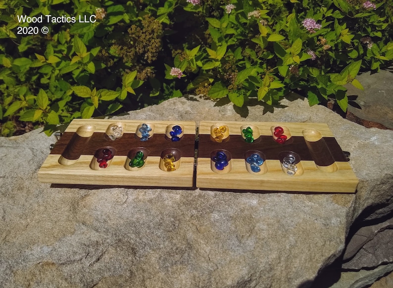 May include: A wooden game board with a walnut inlay and glass marbles in various colours. The board is divided into two sections, each with seven compartments. The game is called "Mancala".