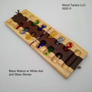 May include: A wooden game board with a walnut and ash wood inlay. The board is divided into two sections, each with a row of holes. The holes are filled with colourful glass stones. The board is closed, and the text "Wood Tactics LLC 2020 &copy;" is visible on the top of the board. The text "Black Walnut w/ White Ash and Glass Stones" is visible at the bottom of the board.