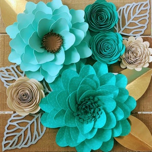 May include: A close-up of a teal and beige paper flower arrangement. The arrangement features several large teal flowers with a layered, ruffled design, along with smaller beige flowers with a spiral design. The flowers are arranged on a wooden background.