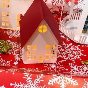 Puede incluir: Una escena de pueblo navideño de papel con una casa de techo rojo, una valla blanca de madera y dos pequeños árboles de Navidad. Las casas tienen ventanas recortadas que dejan pasar la luz.