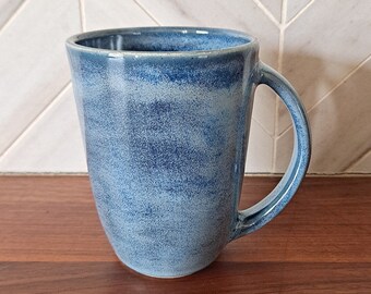May include: A handmade ceramic mug with a blue speckled glaze. The mug has a large handle and a slightly tapered shape. The mug is sitting on a wooden surface.
