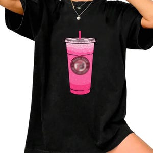 Könnte beinhalten: Schwarzes T-Shirt mit einem rosa Getränk-Motiv. Das Getränk befindet sich in einem transparenten Becher mit rosa Deckel und Strohhalm. Das Motiv enthält ein rosa Logo. Das Shirt ist ein lässiger, übergroßer Stil.