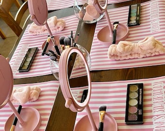 May include: A table setting with pink and white striped placemats, each with a pink vanity mirror, makeup brushes, and an eyeshadow palette. Also includes pink headbands and glass bowls with colorful cotton balls.