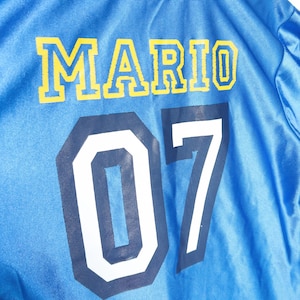 Puede incluir: Una chaqueta de color azul brillante con acabado satinado que presenta el nombre MARIO impreso en letras amarillas llamativas, con el n&uacute;mero 07 debajo en grandes d&iacute;gitos blancos con borde azul oscuro, creando un estilo deportivo.