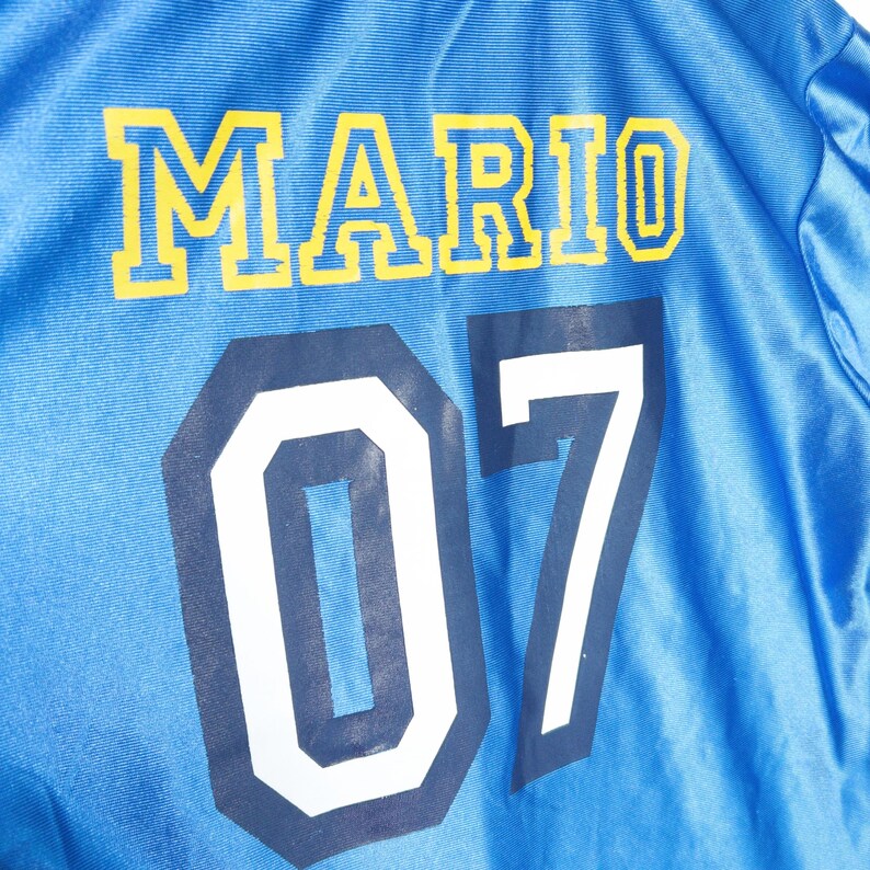 Puede incluir: Una chaqueta de color azul brillante con acabado satinado que presenta el nombre MARIO impreso en letras amarillas llamativas, con el n&uacute;mero 07 debajo en grandes d&iacute;gitos blancos con borde azul oscuro, creando un estilo deportivo.