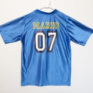 Puede incluir: Una camiseta de manga corta de color azul brillante con el nombre MARIO en letras amarillas en la parte superior de la espalda. Debajo del nombre, el n&uacute;mero 07 est&aacute; impreso en grandes d&iacute;gitos azul marino oscuro con un borde blanco.