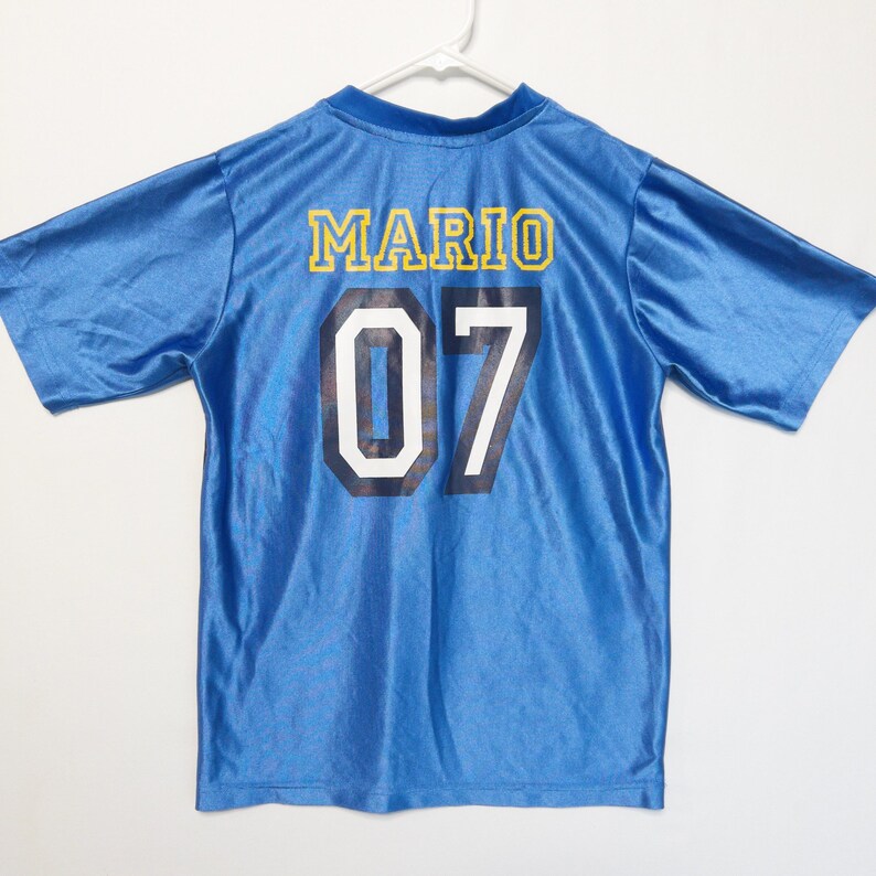 Puede incluir: Una camiseta de manga corta de color azul brillante con el nombre MARIO en letras amarillas en la parte superior de la espalda. Debajo del nombre, el n&uacute;mero 07 est&aacute; impreso en grandes d&iacute;gitos azul marino oscuro con un borde blanco.