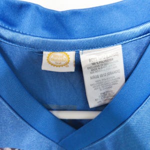 Puede incluir: Una camiseta azul con cuello en V que presenta una etiqueta Official Nintendo Seal y una etiqueta de cuidado blanca. La etiqueta especifica 100% poli&eacute;ster, talla 10/12 Large, e indica que la prenda fue fabricada en Bangladesh. Textura sint&eacute;tica lisa.